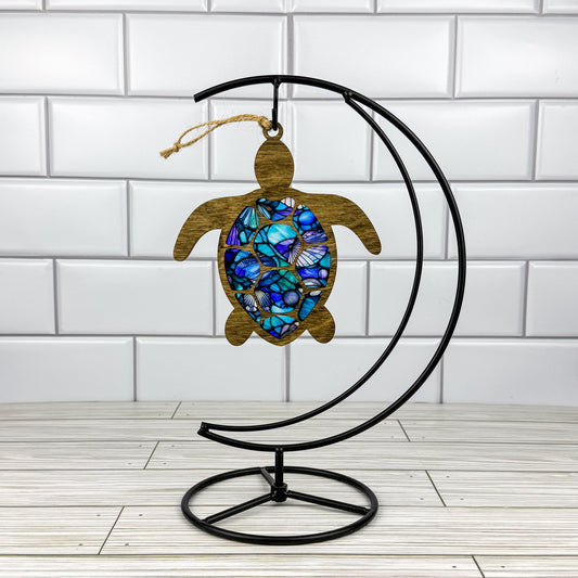 Sea turtle suncatcher made from wood and acrylic hanging on a metal stand in front of a white tiled wall.