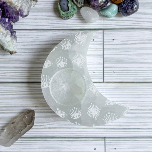 Crescent moon shaped selenite candle holder with engraved eye patterns on a wooden surface with crystals around