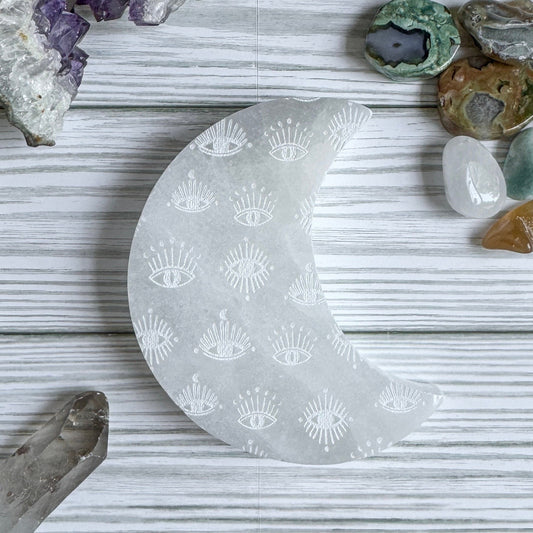 Selenite crescent moon charging plate with engraved eye patterns on a textured surface with crystals around
