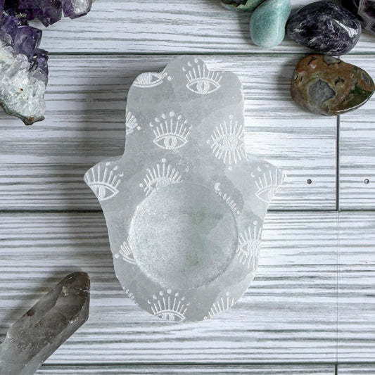 Hamsa shaped selenite candle holder with engraved eye patterns on a wooden surface with crystals around