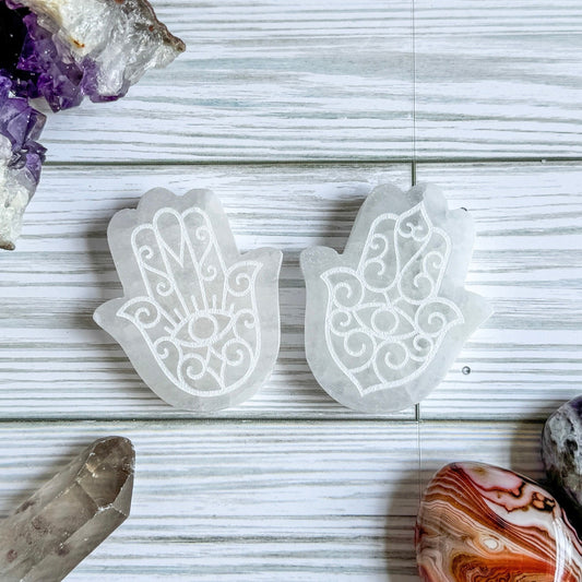 Two engraved white selenite Hamsa hand stones on a light wooden surface with crystals around.