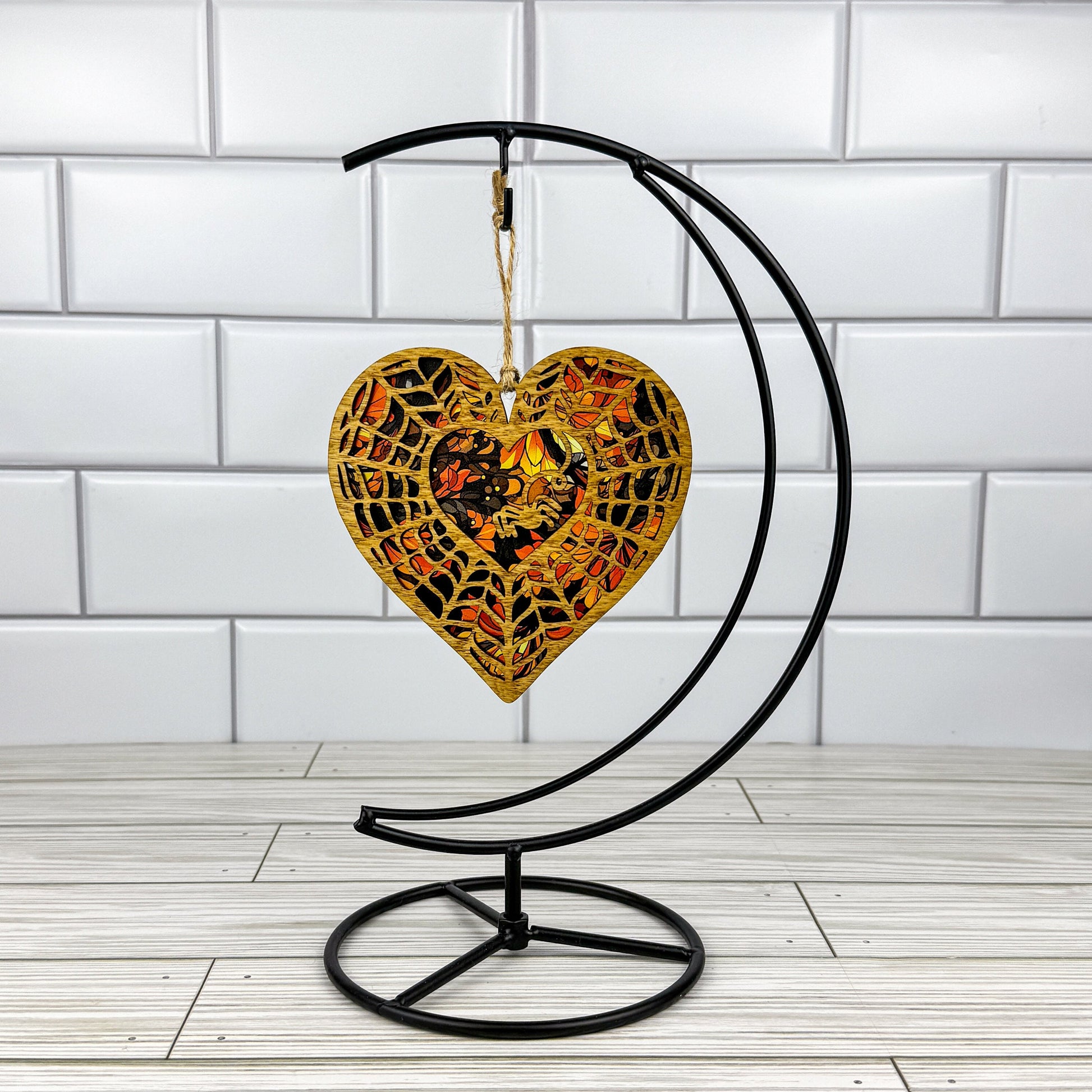 Spiderweb heart suncatcher made from wood and acrylic hanging on a metal stand in front of a white tiled wall.