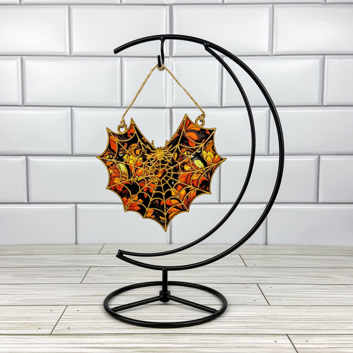 Spiderweb suncatcher made from wood and acrylic hanging on a metal stand in front of a white tiled wall.