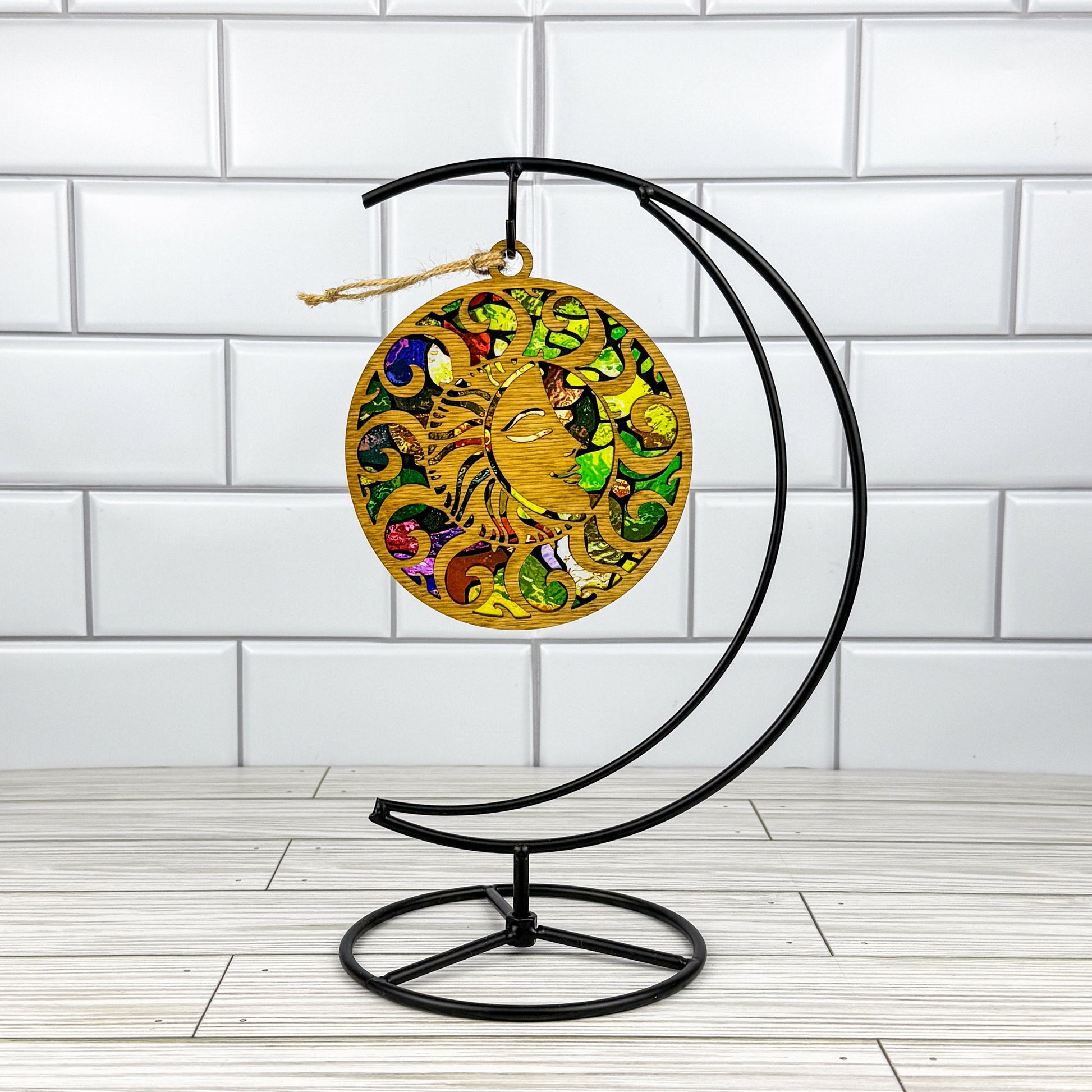 Sun shaped suncatcher made from wood and acrylic hanging on a metal stand in front of a white tiled wall.