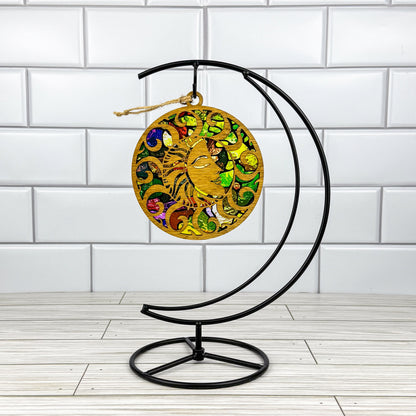 Sun shaped suncatcher made from wood and acrylic hanging on a metal stand in front of a white tiled wall.